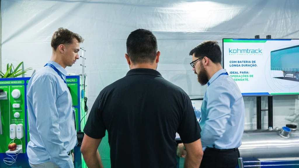 Dois homens de camisa azul-clara atendem visitante de camisa polo preta entre o painel de sensores wireless e uma TV que exibe a Kohmtrack