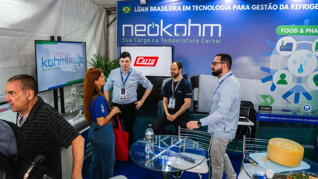 Quatro pessoas conversam no interior do estande da Neokohm na FENATRAN 2024, com TV exibindo a Kohmtrack e painel Food & Pharma ao fundo.