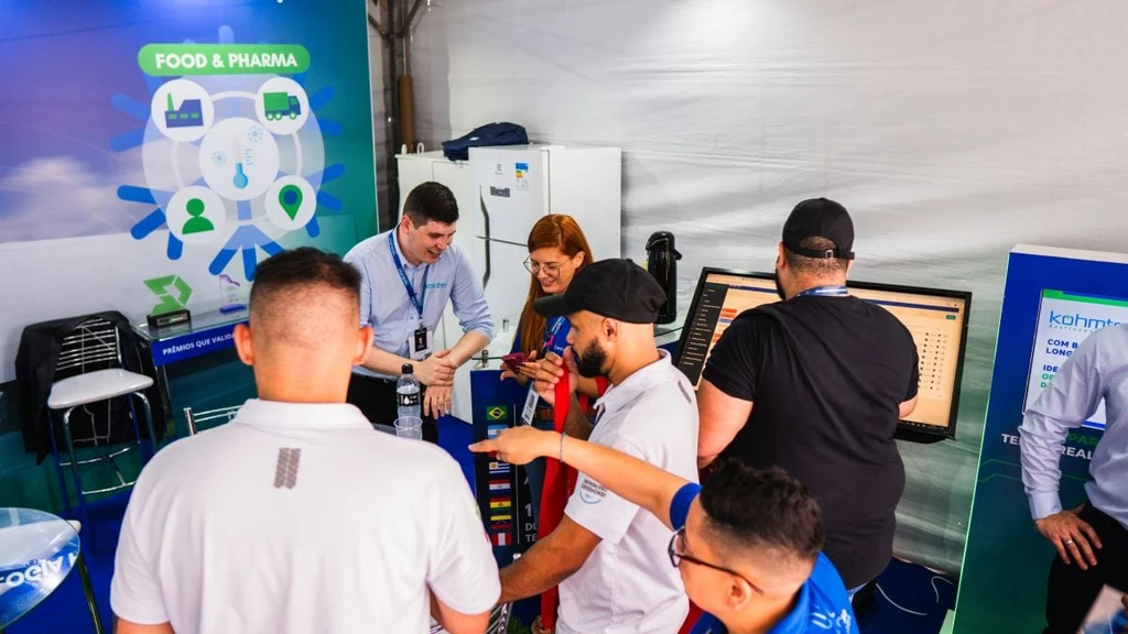 Visto de cima, grupo de visitantes de boné e camisa polo se reúne no balcão, onde homem de camisa azul-clara sorri e outro olha um monitor