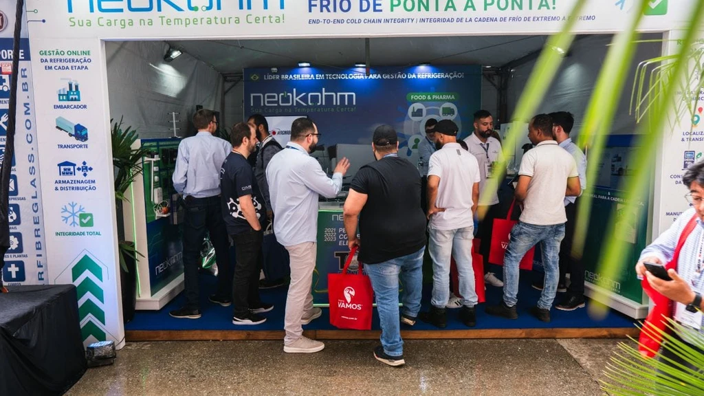 Vista frontal do estande da Neokohm lotado, com visitantes de sacolas vermelhas junto ao balcão e fachada com “Sua Carga na Temperatura Certa!”