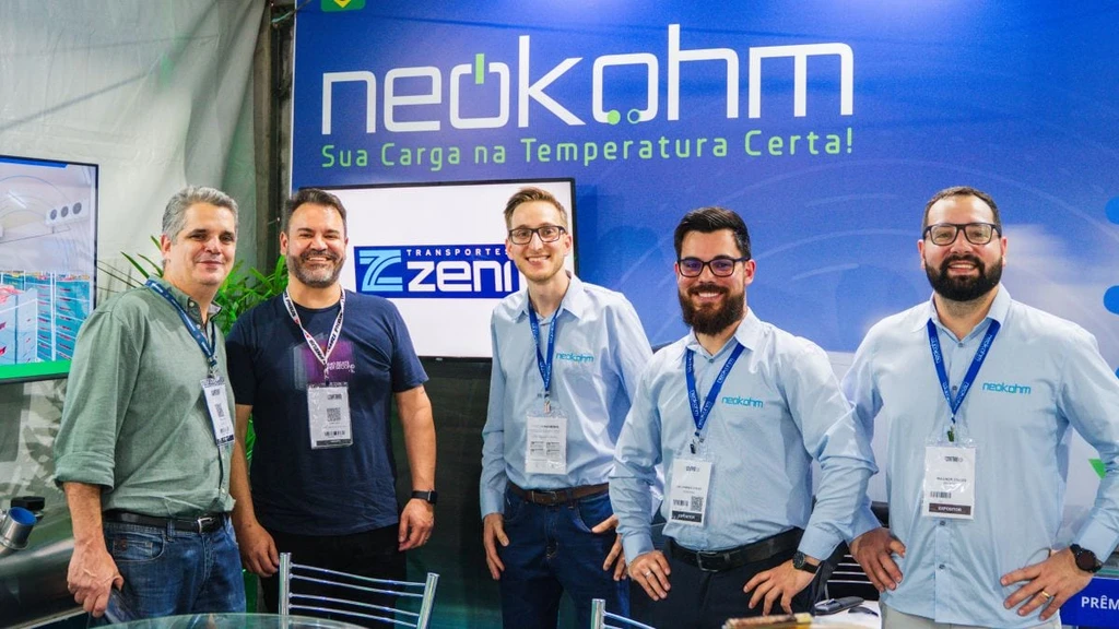 Homem de camisa verde e outro de camiseta azul-marinho posam com três homens de camisa azul-clara da Neokohm diante do painel da marca