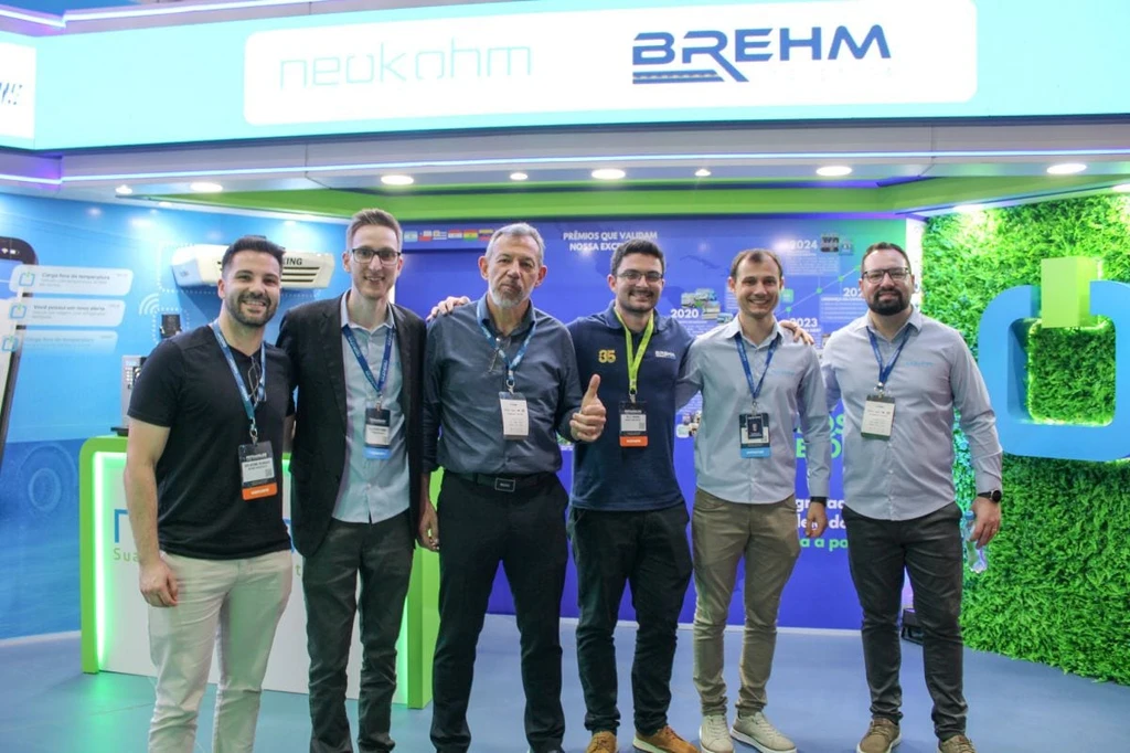 Seis homens com crachás posam sob painel com os logotipos da Neokohm e da Brehm; um deles, de cabelo grisalho, faz sinal de positivo