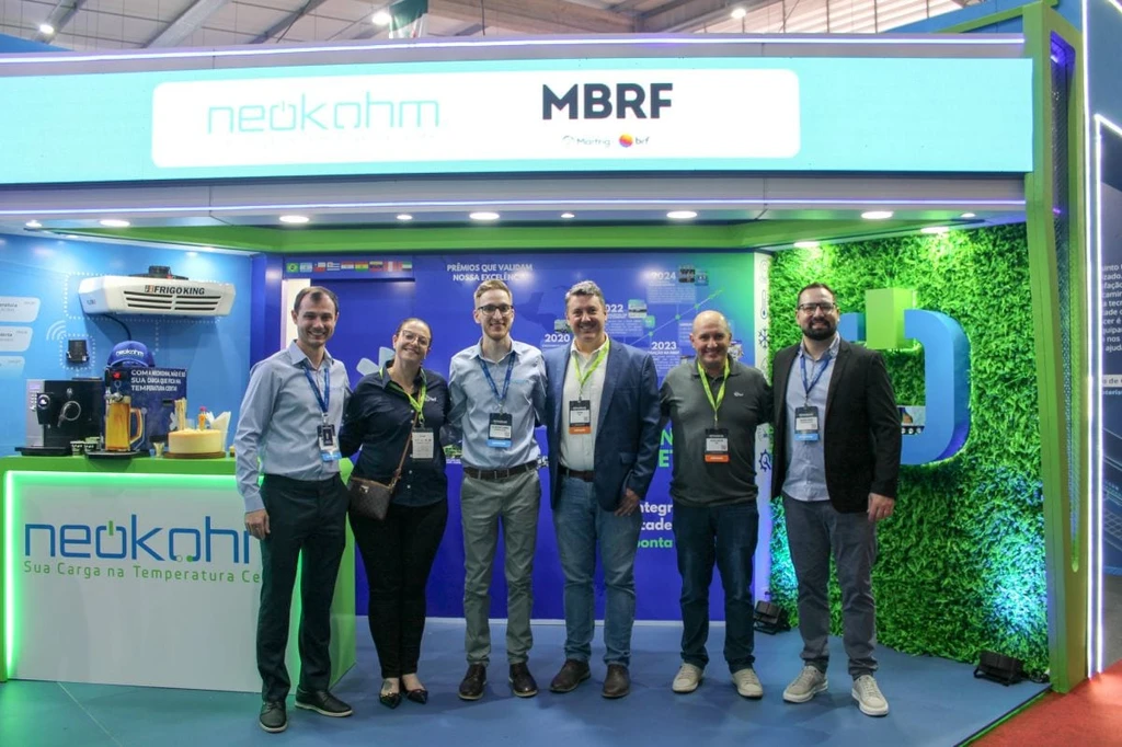 Cinco homens e uma mulher com crachás posam entre o balcão e a parede verde do estande, sob painel com os logotipos da Neokohm e da MBRF