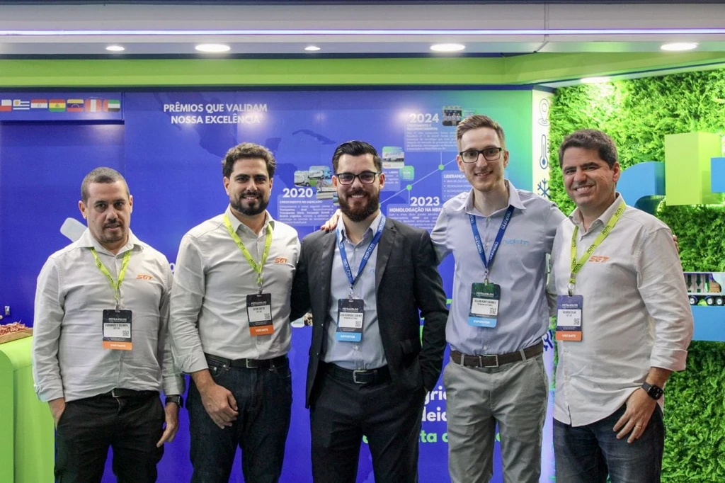Cinco homens com crachás posam lado a lado diante de painel azul com a frase “Prêmios que validam nossa excelência” e uma linha do tempo