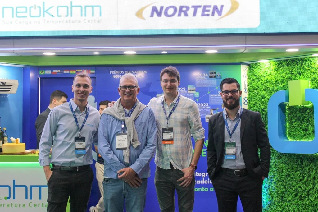 Quatro homens com crachás posam sob painel com os logotipos da Neokohm e da Norten; um deles, de cabelo grisalho, tem um suéter sobre os ombros