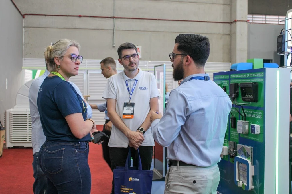 Homem de camisa azul-clara conversa com visitantes ao lado de painel de telemetria online da refrigeração, com dispositivos e sensores, no estande da Neokohm