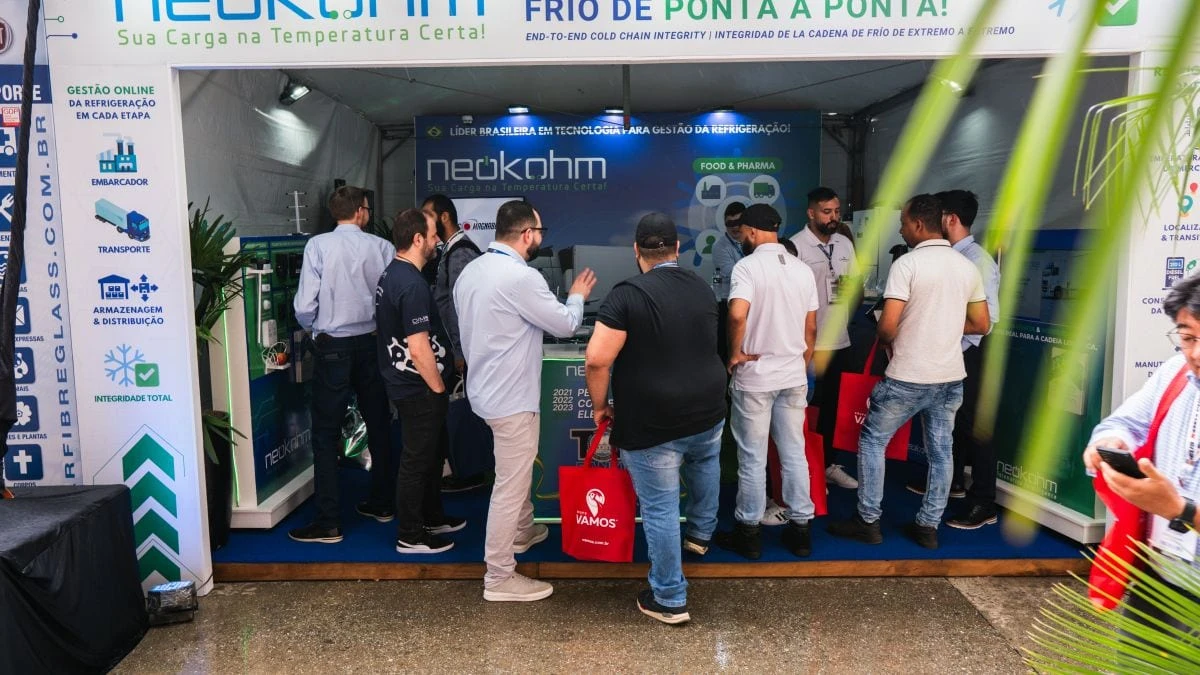 Vista frontal do estande da Neokohm lotado, com visitantes de sacolas vermelhas junto ao balcão e fachada com “Sua Carga na Temperatura Certa!”