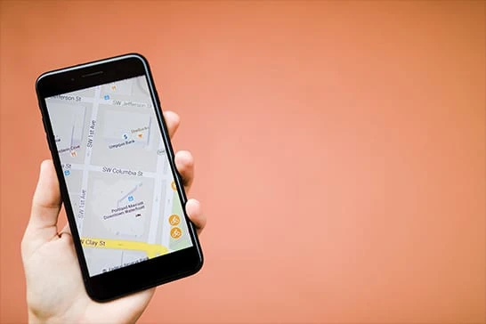 Mão segurando um smartphone com um aplicativo de mapa de ruas aberto na tela, diante de um fundo liso laranja-salmão desfocado.