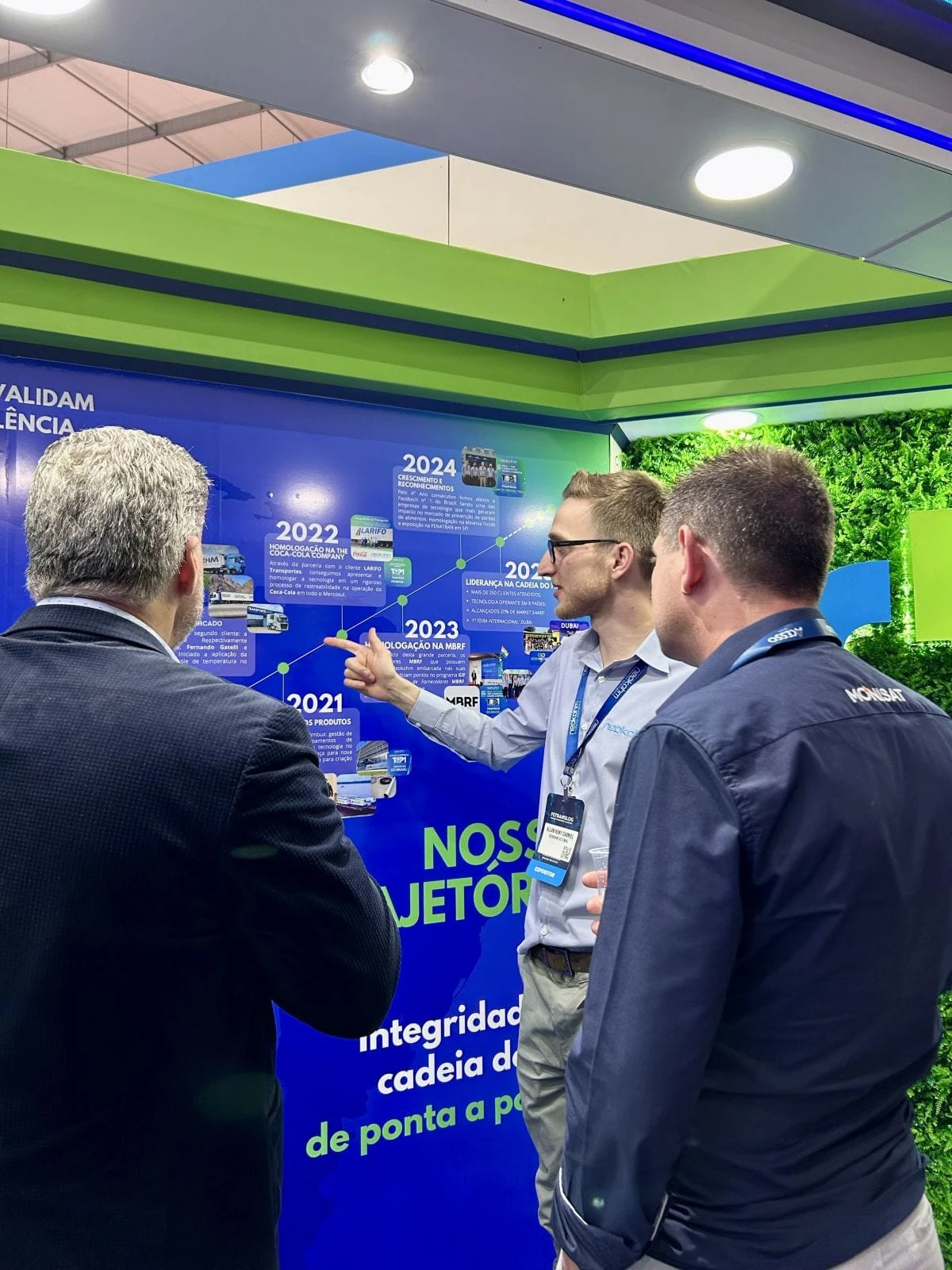 Integrante da Neokohm aponta o painel “Nossa trajetória” enquanto conversa com dois visitantes no estande da Fetranslog 2025.