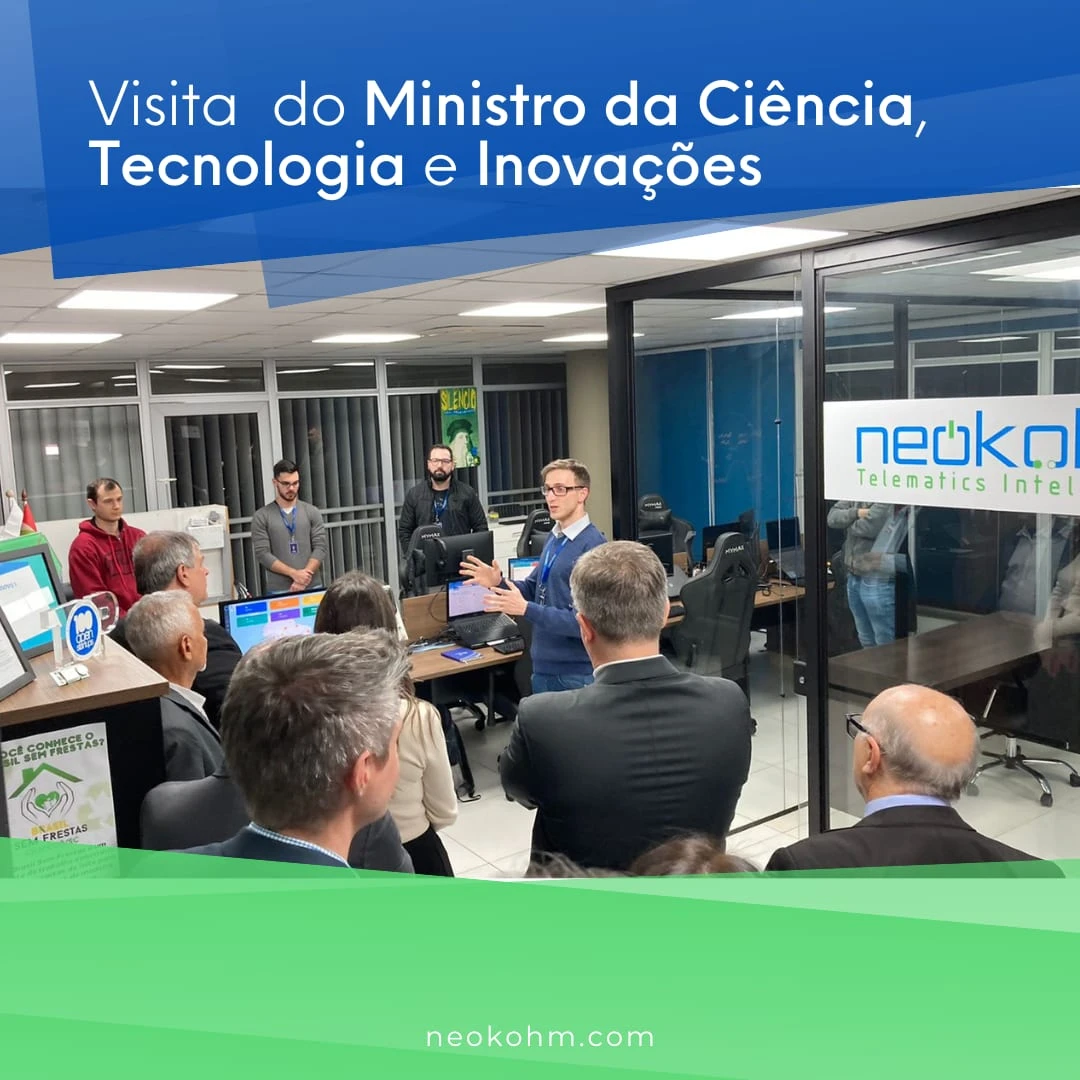 Homem de óculos e suéter azul gesticula ao falar com visitantes de terno no escritório, ao lado de divisória de vidro com o logotipo da Neokohm.