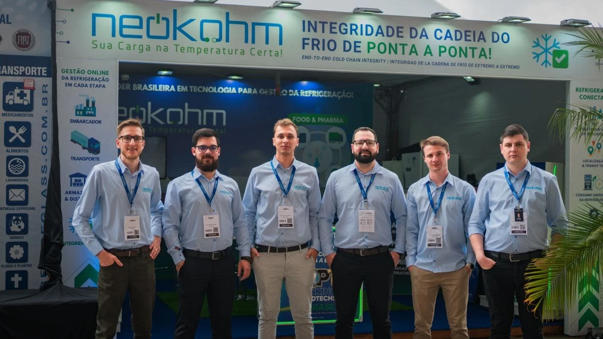 Seis integrantes da equipe Neokohm, de camisa azul e crachá, posam em frente ao estande da empresa na FENATRAN 2024