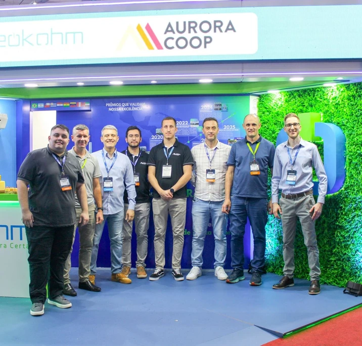 Oito homens com crachás posam lado a lado no estande, sob painel com os logotipos da Neokohm e da Aurora Coop