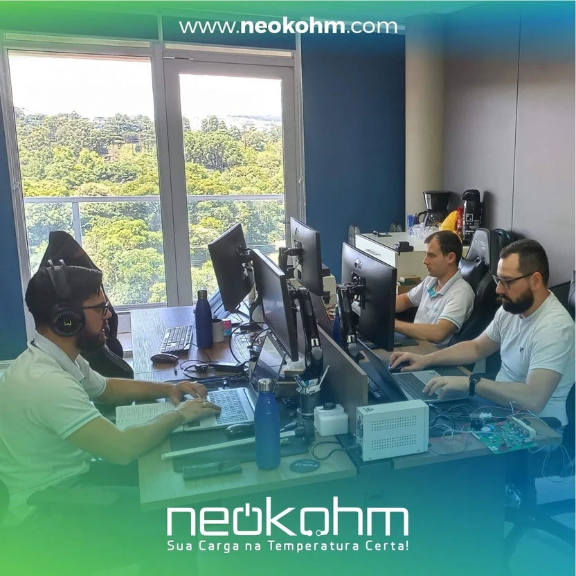 Três homens trabalham em notebooks e monitores na sala de Pesquisa e Desenvolvimento de Produto da Neokohm, com placa eletrônica sobre a bancada.