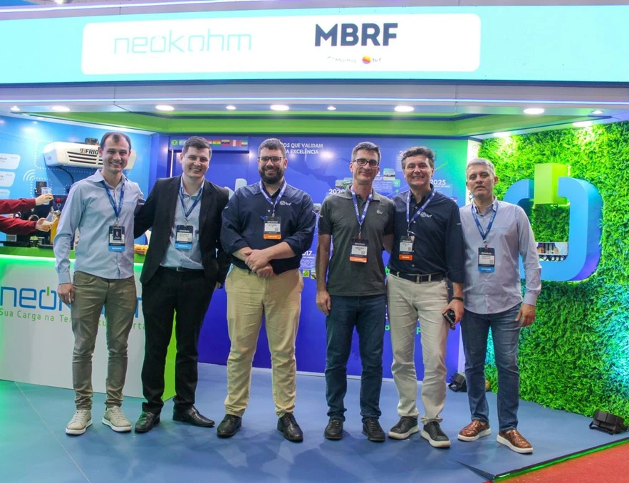 Seis homens com crachás posam sob painel com os logotipos da Neokohm e da MBRF; à esquerda, uma mão segura um copo sob a torneira do balcão